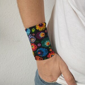 Pu&ograve; includere: Un braccialetto nero per il polso con un motivo floreale colorato. Il motivo presenta colori vivaci come rosso, giallo, blu, verde e viola. Il testo "Folklore Black" &egrave; visibile nella parte inferiore dell'immagine.