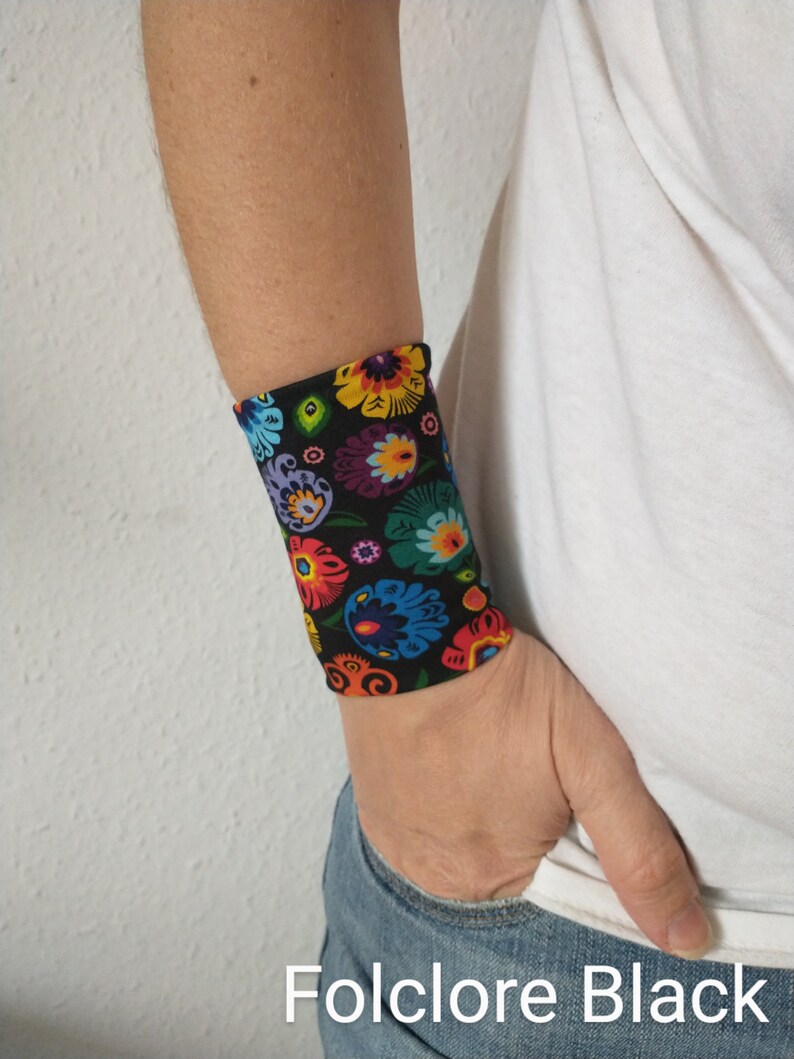 Pu&ograve; includere: Un braccialetto nero per il polso con un motivo floreale colorato. Il motivo presenta colori vivaci come rosso, giallo, blu, verde e viola. Il testo "Folklore Black" &egrave; visibile nella parte inferiore dell'immagine.