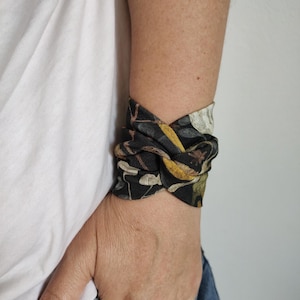 May include: A patterned fabric wrist wrap with the text "Golden Leaves". The wrap, knotted on the wrist, features a black base with gold, yellow, and white floral details. The wrist wrap is a stylish accessory.