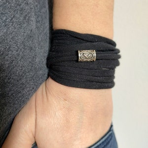 Op de afbeelding: Een zwarte stoffen armband met een zilveren bedel met een geometrisch ontwerp.
