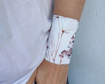 Può includere: Un braccialetto da polso in tessuto bianco con un motivo floreale di dente di leone marroni e viola. Il testo "Dente di leone" è stampato sotto il braccialetto.