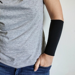 Op de afbeelding: Een persoon draagt een grijs t-shirt en een blauwe jeans met een zwarte armbeschermer die hun onderarm bedekt.