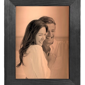 May include: A black wood picture frame with a sepia toned photo of a couple smiling and hugging. The photo is set against a peach colored background.