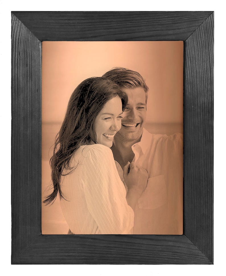 May include: A black wood picture frame with a sepia toned photo of a couple smiling and hugging. The photo is set against a peach colored background.