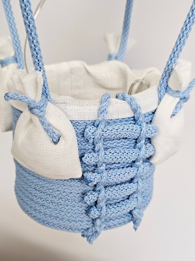 Puede incluir: Un macetero colgante azul claro hecho de cuerda trenzada con un forro de tela blanca. El macetero tiene dos peque&ntilde;as bolsas de tela blanca unidas a los lados.