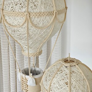 May include: Two white string-wrapped hot air balloon lamps hanging from the ceiling. The larger balloon has a basket made of string and a white fabric lining. The smaller balloon is hanging below the larger balloon.
