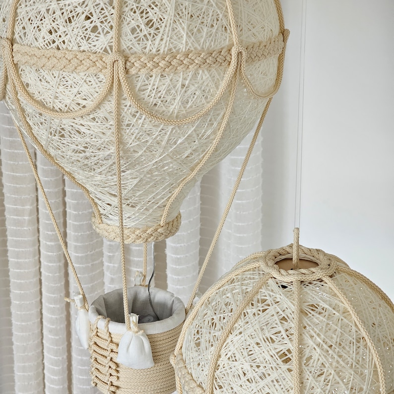 May include: Two white string-wrapped hot air balloon lamps hanging from the ceiling. The larger balloon has a basket made of string and a white fabric lining. The smaller balloon is hanging below the larger balloon.