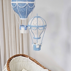 Puede incluir: Dos globos aerost&aacute;ticos de cuerda azul y blanco colgando sobre una cuna de mimbre con un colch&oacute;n blanco y ribete de encaje.