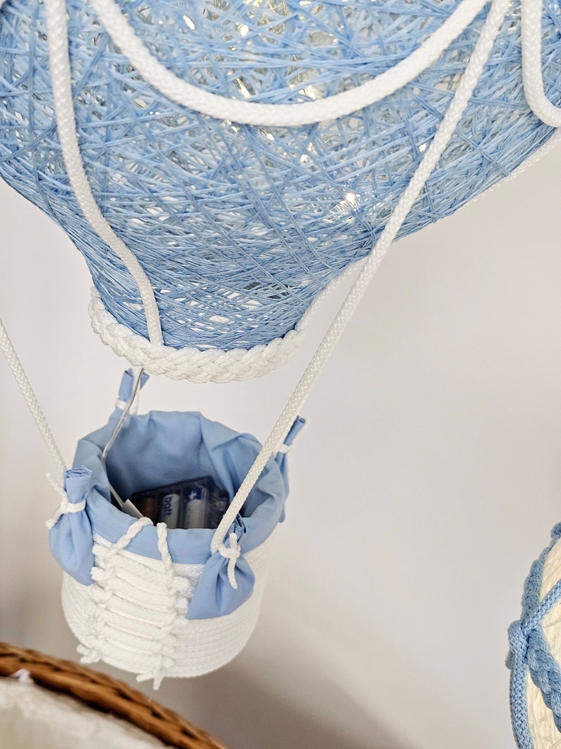 Puede incluir: Un globo aerost&aacute;tico de luz de cuerda azul y blanco con una cesta de tela blanca. La cesta tiene un forro de tela azul y contiene bater&iacute;as.