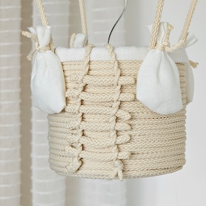 May include: A white, crocheted hot air balloon with a white fabric lining and two small white fabric pouches hanging from the sides. The balloon is suspended from a white ceiling.