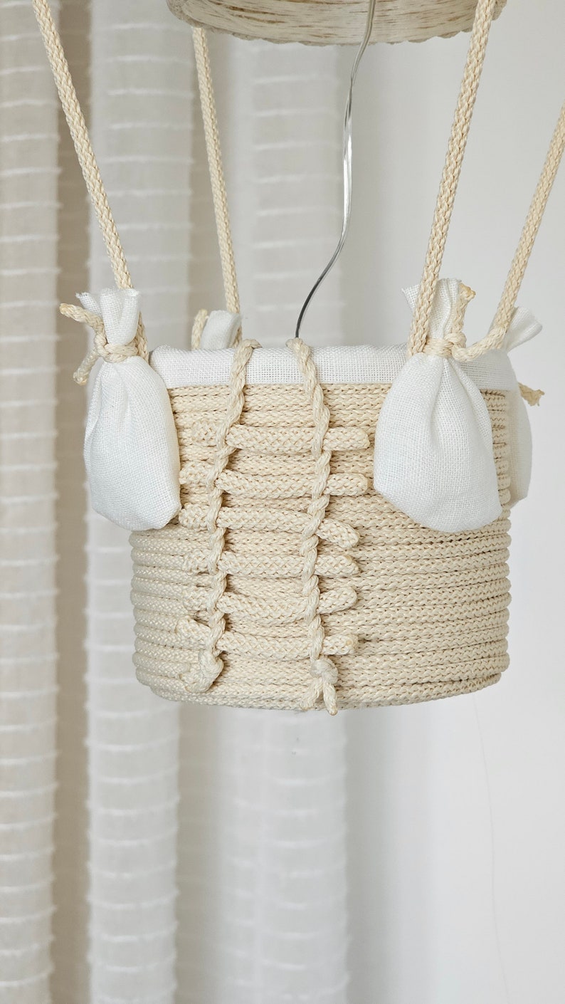 May include: A white, crocheted hot air balloon with a white fabric lining and two small white fabric pouches hanging from the sides. The balloon is suspended from a white ceiling.
