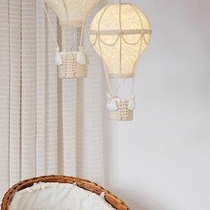 May include: Two white, string-wrapped hot air balloons with lights inside hang from the ceiling above a wicker bassinet with a white lining.