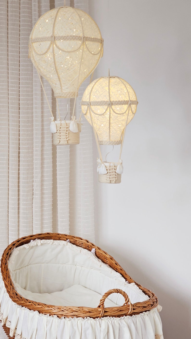 May include: Two white, string-wrapped hot air balloons with lights inside hang from the ceiling above a wicker bassinet with a white lining.