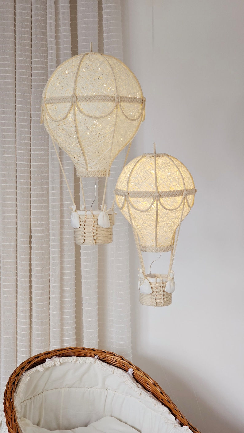 May include: Two white, string-wrapped hot air balloons with lights inside, hanging from the ceiling above a white wicker bassinet.