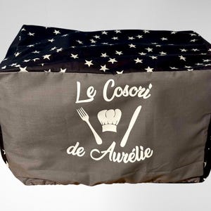 Peut inclure: Une housse en tissu gris et noir avec des étoiles blanches et le texte "Le Cosori de Aurélie" avec un chapeau de chef et une fourchette et un couteau.