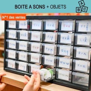 Könnte beinhalten: Ein schwarzer Kunststoff-Aufbewahrungsschrank mit mehreren durchsichtigen Schubladen, die jeweils mit einer weißen Karte beschriftet sind, auf der ein Buchstabe des Alphabets in Schreibschrift steht. Oben steht BOITE A SONS + OBJETS mit ABC-Symbol.