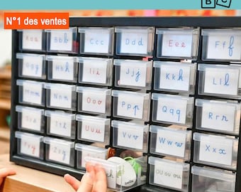 May include: A black plastic storage cabinet with multiple clear drawers, each labeled with a white card featuring a letter of the alphabet in cursive script. The text BOITE A SONS + OBJETS and an ABC block icon appear at the top of the image.