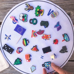 Könnte beinhalten: Eine runde weiße Matte mit blauem Rand, auf der verschiedene bunte Schuhanhänger verstreut sind, darunter eine Flagge der Europäischen Union, ein Papagei, ein Domino, ein Lagerfeuer, ein Roboter, eine Palme, ein Gewicht, ein Zug und ein Tannenbaum.