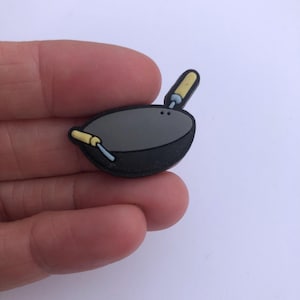 May include: A small, cartoon-style charm in the shape of a wok, featuring a gray cooking surface and a black rim. Two yellow and blue cooking utensils are included. The charm is held in a hand.