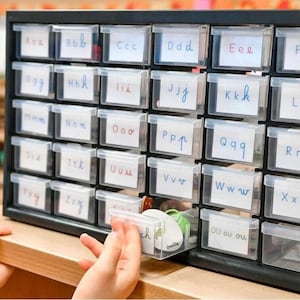 Puede incluir: Un mueble de almacenamiento negro con múltiples cajones de plástico transparente, cada uno etiquetado con una letra del alfabeto en escritura manuscrita. Las manos de un niño interactúan con el cajón. La unidad está sobre una superficie de madera.