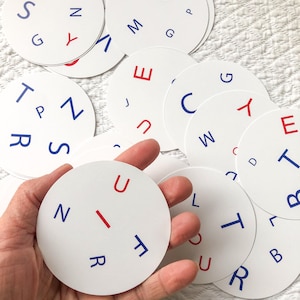 Peut inclure: Plusieurs cartes circulaires blanches avec des lettres rouges et bleues. Une carte est tenue dans une main. Les lettres sont disposées autour du périmètre des cercles.