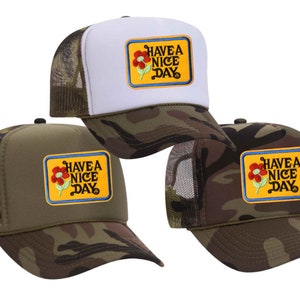 Op de afbeelding: Drie trucker caps met een camouflagepatroon en een geel patch met de tekst "Have a Nice Day" met een rode bloem afbeelding.