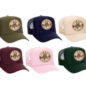 May include: Six trucker hats in various colors: olive green, navy blue, beige, burgundy, pink, and dark green. Each hat features a circular patch with the text "WHAT IF IT ALL WORKS OUT?" and a floral design.