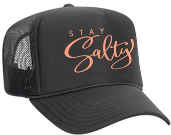 May include: Black trucker hat with a mesh back. The front of the hat has the text "Stay Salty" in a peach colored script font.