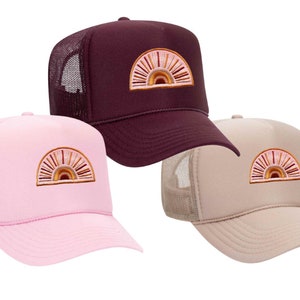May include: Three trucker hats with a rainbow sun design. The hats are in pink, burgundy, and beige.