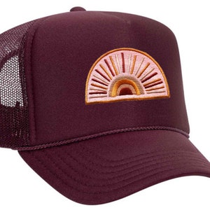 Peut inclure: Un chapeau de camionneur bordeaux avec un motif soleil brodé rose et orange. Le chapeau a un panneau arrière en maille.