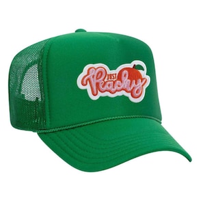 May include: A green trucker hat with a mesh back and a curved brim. The hat features a patch with the words "Just Peachy" in white script, along with a peach illustration in pink and orange.