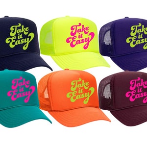 May include: Six trucker hats with mesh back panels. The hats are in different colors: navy blue, neon yellow, purple, teal, orange, and maroon. Each hat has the phrase "Take it Easy" written in a retro font in pink or yellow.
