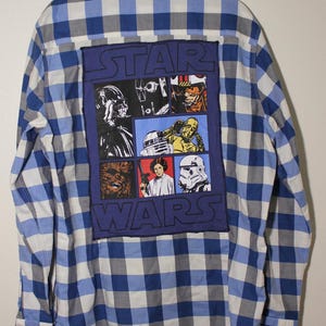 May include: A blue and white plaid long-sleeved shirt with a Star Wars patch on the back. The patch features a collage of characters from the Star Wars franchise, including Darth Vader, Chewbacca, Princess Leia, and a Stormtrooper. The text "STAR WARS" is printed in large letters above the characters.
