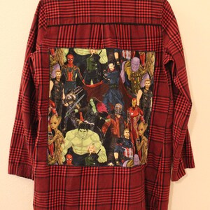 May include: A red and black plaid flannel shirt with a large patch on the back featuring a collage of Marvel superhero characters, including Iron Man, Captain America, Thor, Hulk, Spider-Man, and more.