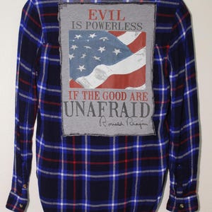 May include: A blue and red checked flannel shirt with a grey patch on the back. The patch features an American flag and the text "Evil is powerless if the good are unafraid." with Ronald Reagan's signature.