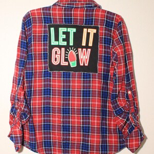 May include: Red and blue checked flannel shirt with a black patch that says "LET IT GLOW" in colourful letters with a light bulb graphic.