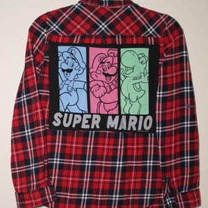 May include: Red and black plaid flannel shirt with a Super Mario Bros. graphic on the back. The graphic features Mario, Luigi, and Yoshi in colorful squares.