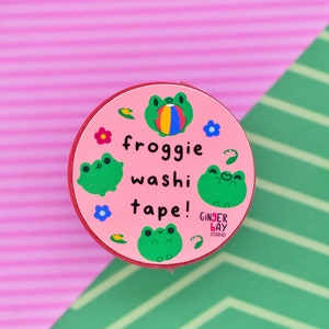 May include: Pink round washi tape dispenser with a frog design. The dispenser has the text "froggy washi tape!" and a small logo that says "Ginger Bay Studios".