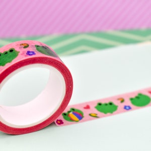 May include: Pink washi tape with green frogs, flowers, and hearts on a white background.