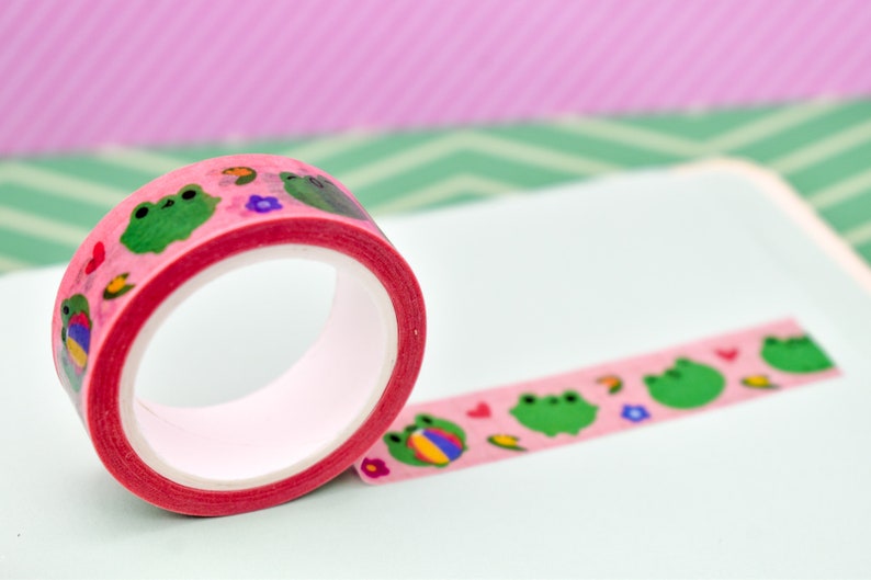 May include: Pink washi tape with green frogs, flowers, and hearts on a white background.