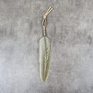 Pode incluir: Um ornamento em forma de pena na cor verde sálvia com uma nervura central e superfície texturizada. O objeto está preso a uma alça de cordão de couro bege claro no topo, fixada com um pequeno anel metálico para pendurar.