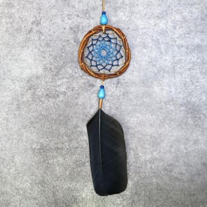 May include: A dream catcher featuring a circular brown twig frame with a woven blue and white web pattern. The ornament includes two light blue beads, one at the top and one at the bottom, and a single large black feather hanging from the base.