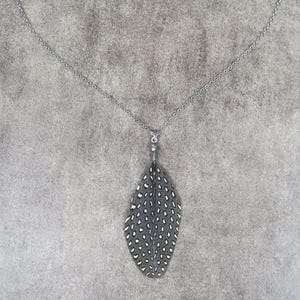 Könnte beinhalten: Eine Halskette mit einer dunklen Metallkette und einem Anhänger in Form einer einzelnen Feder. Die Feder ist dunkelgrau und mit einem Muster aus kleinen, weißen, gleichmäßig verteilten Punkten versehen, befestigt an einer kleinen Metallöse.