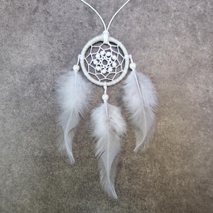 Peut inclure: Un attrape-rêves blanc avec un cadre circulaire tissé, orné de perles blanches et de trois grandes plumes blanches et duveteuses. L'attrape-rêves est suspendu à un cordon blanc sur un fond gris.