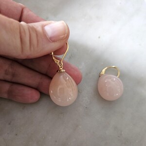 Peut inclure: Une paire de boucles d'oreilles avec des fermoirs dorés et des pierres en forme de goutte d'eau de couleur pêche. Une boucle d'oreille est tenue par une main, l'autre est posée sur une surface claire.