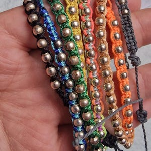 Könnte beinhalten: Eine Sammlung von sechs verstellbaren Armbändern mit metallisch silberfarbenen Perlen, die in bunte Nylonschnüre eingewebt sind. Die Armbänder umfassen schwarze, blaue, grüne, gelbe, rosa und orangefarbene Stränge mit Schiebeknoten.