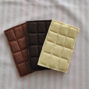 Op de afbeelding: Drie vierkante stukken vilt in de kleuren bruin, zwart en crème, elk met een gestikt patroon van vierkanten.