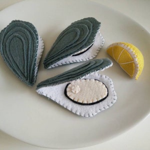 May include: A white plate holds three felt oyster shells in shades of gray, with white interiors and black accents. A felt lemon wedge with white stitching sits nearby. The handmade food toys are designed for play.