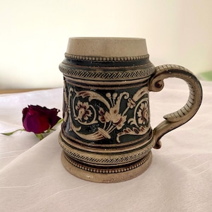 May include: A brown and green ceramic beer stein with a floral design. The stein has a handle and a lid.