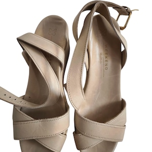 May include: A pair of light beige leather wedge sandals. The sandals feature crisscross straps over the toes and ankle straps with gold-colored buckles. The footbed is stamped with a butterfly logo and the text "WEEKEND MaxMara".